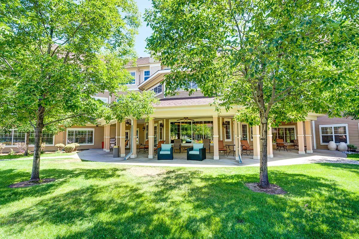 A sunny day view of a building with a covered patio, surrounded by green trees and a well-maintained lawn.