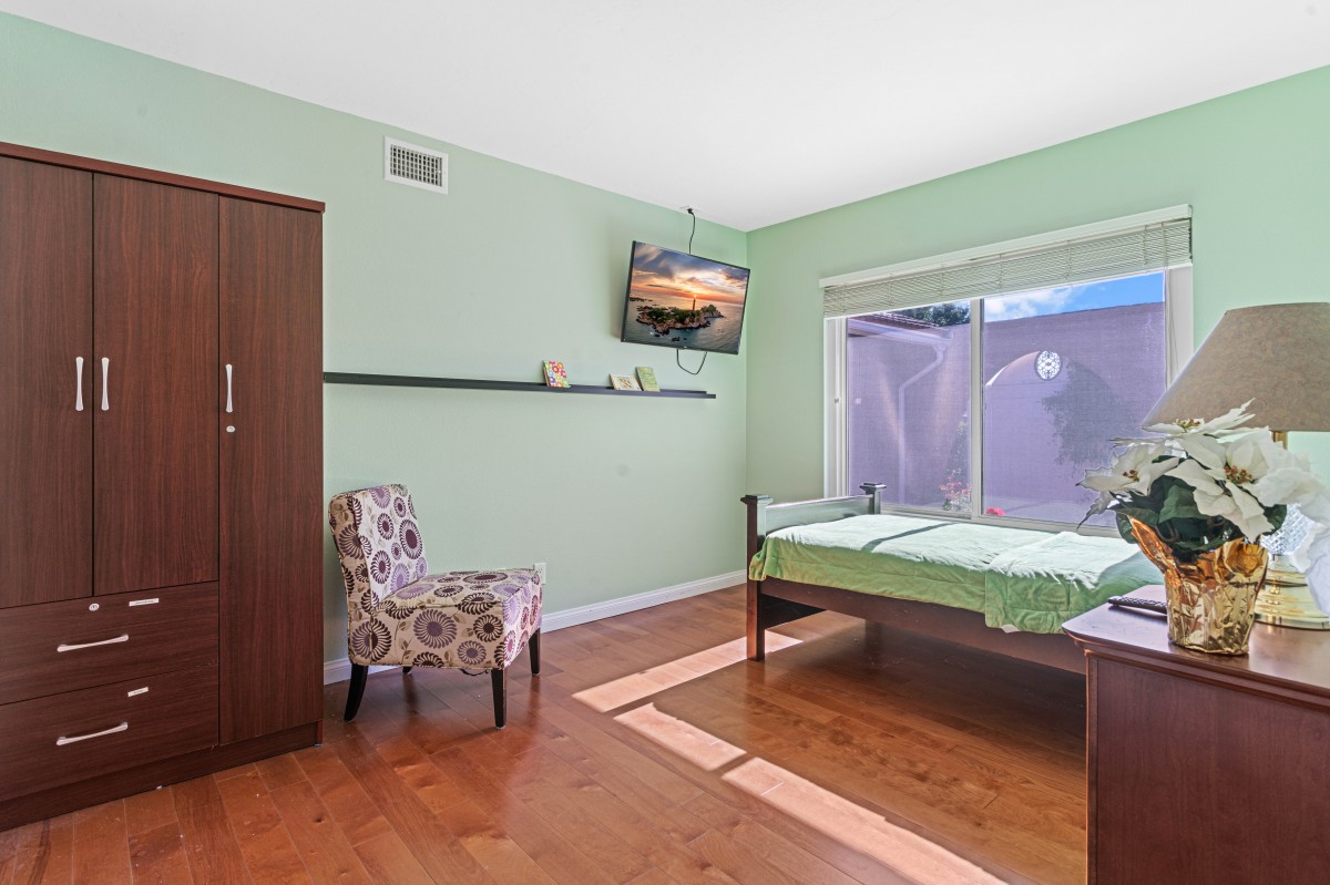 A cozy bedroom with a single bed, green walls, wooden flooring, a floral chair, and a mounted television.