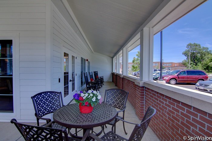 A cozy porch area with a table, chairs, and potted flowers, overlooking a parking lot.