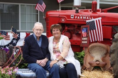 An elderly couple sitting in front of a red Farmall tractor decorated with American flags.