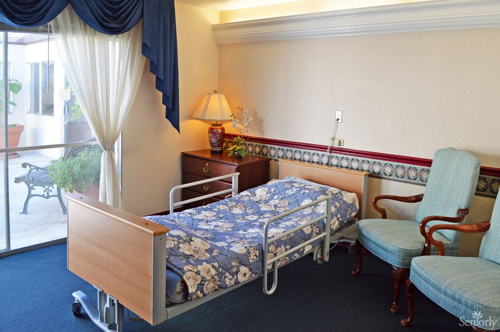 A hospital room featuring a neatly made bed with floral covers, a bedside table with a lamp, and a chair by the window.