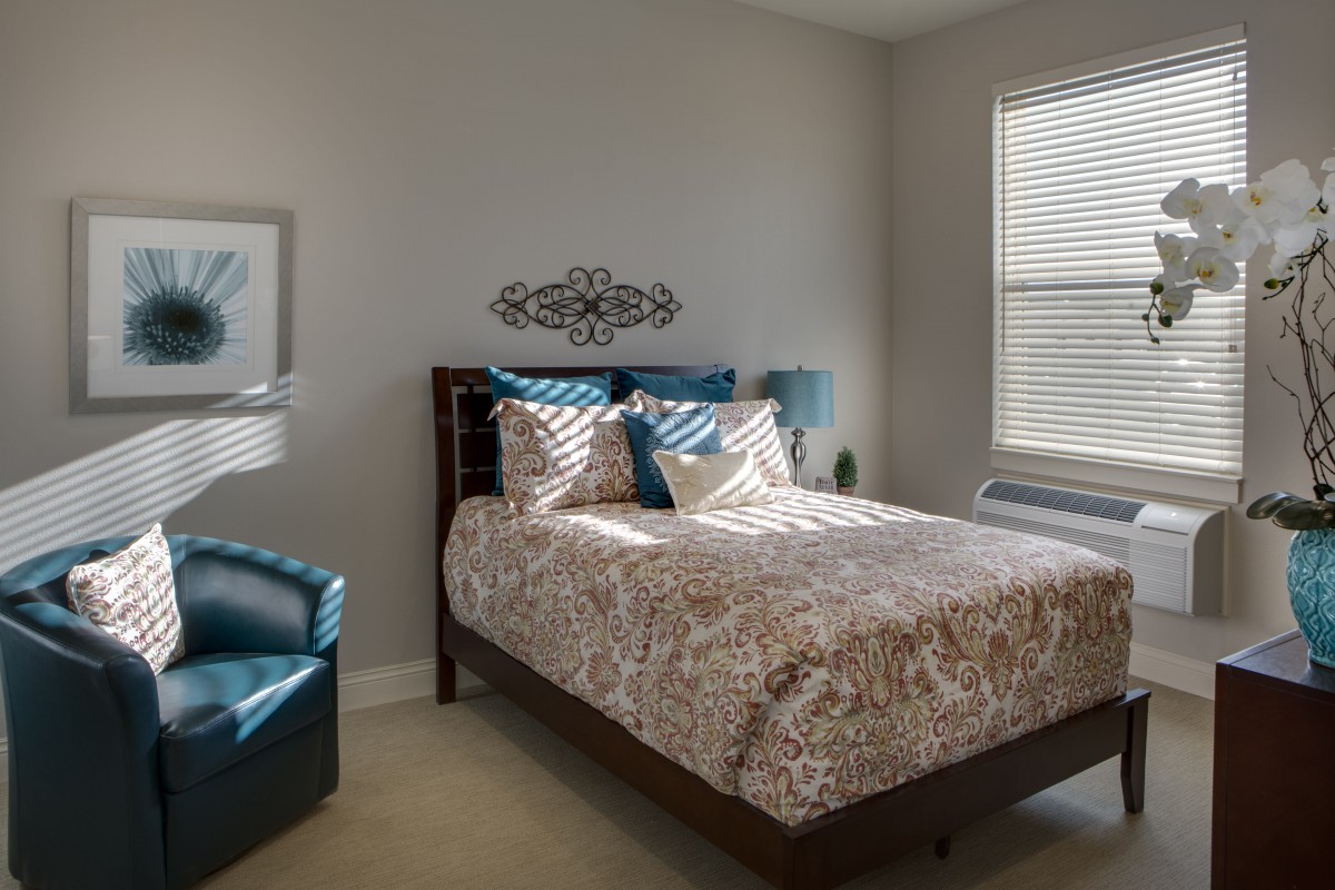 A cozy bedroom with a patterned bedspread, teal chair, and sunlight streaming through the blinds.
