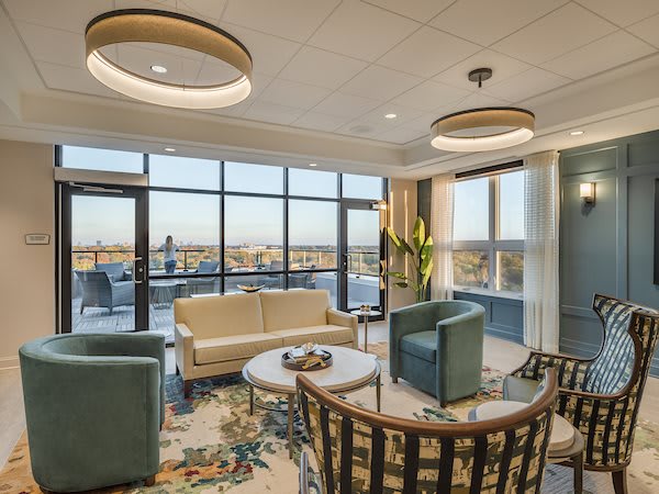 A modern living room with large windows offering a city view, featuring comfortable seating and stylish lighting fixtures.