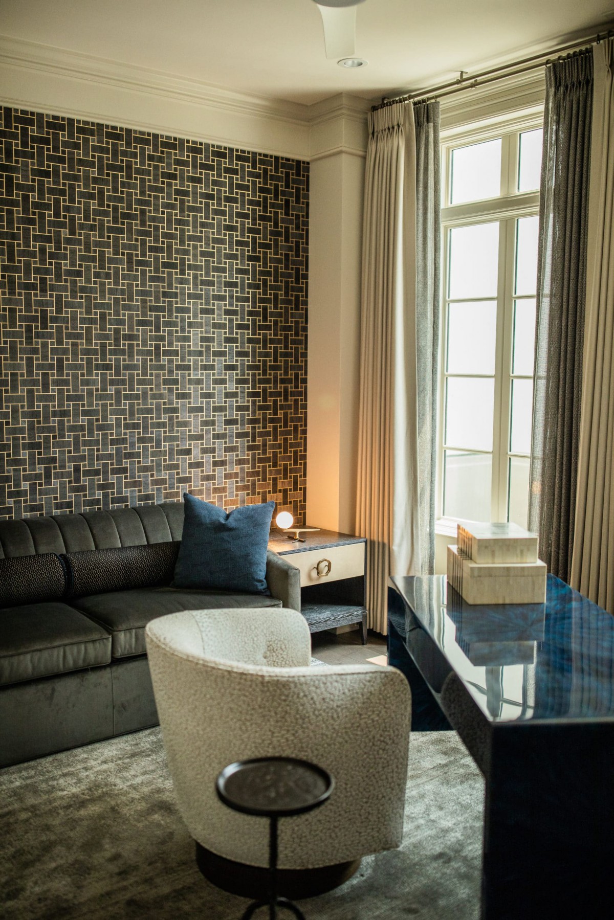 A modern living room featuring a sectional sofa, geometric wallpaper, and ample natural light from large windows.
