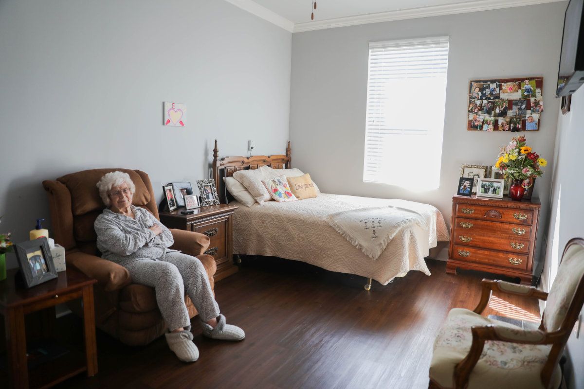 An elderly woman sitting in a chair in a neatly arranged bedroom with wooden furniture and natural light coming through the window.