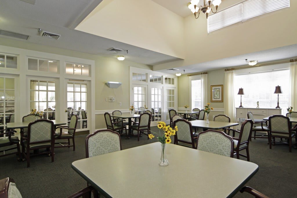 A spacious dining room with neatly arranged tables and chairs, featuring a vase of flowers on the table and a chandelier overhead.