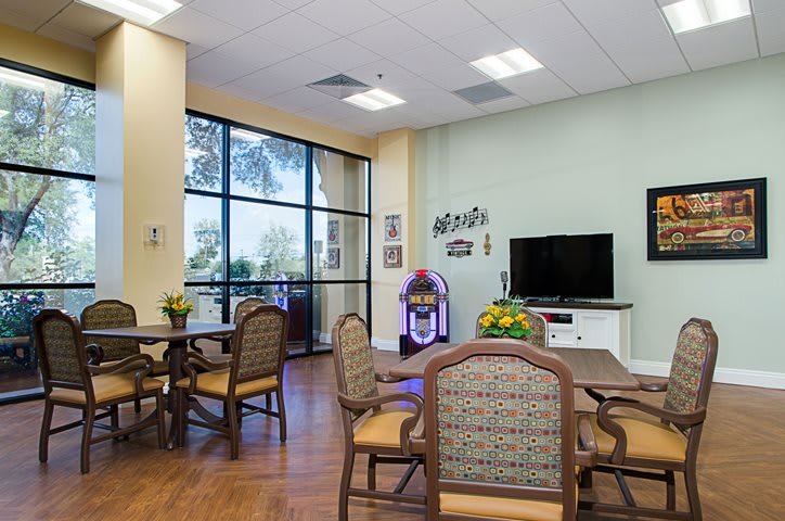 A cozy community room featuring tables, chairs, a television, and a jukebox with large windows providing natural light.