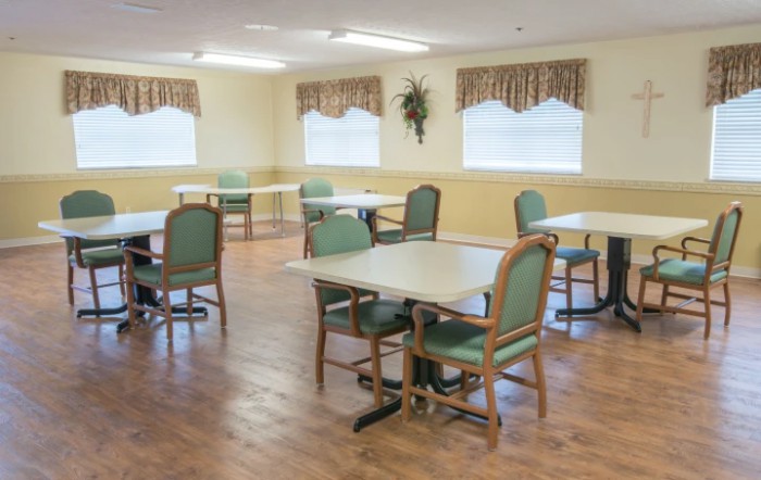 An empty dining area with multiple tables and chairs, featuring a wooden floor and windows with curtains.