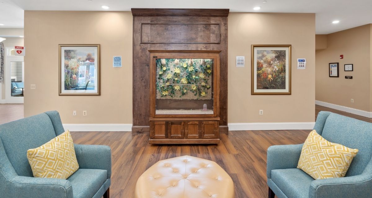 A cozy waiting room featuring an aquarium set within a wooden paneled wall, surrounded by artwork and comfortable seating.