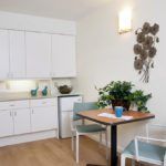 A compact kitchen and dining area with white cabinets, a small fridge, and a wooden table adorned with an indoor plant and modern wall art.