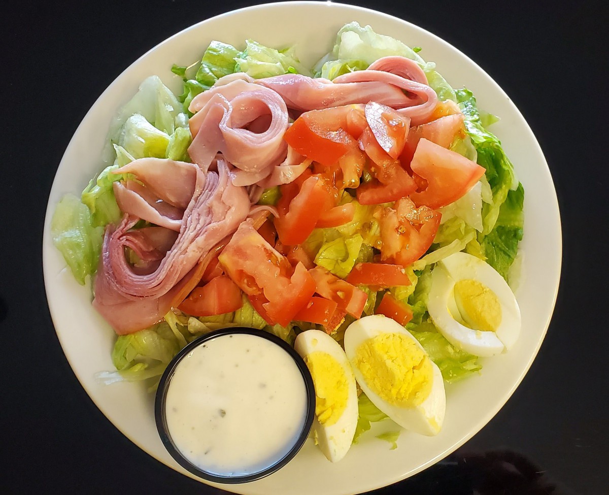 A fresh salad with ham, tomatoes, lettuce, hard-boiled eggs, and a side of dressing.
