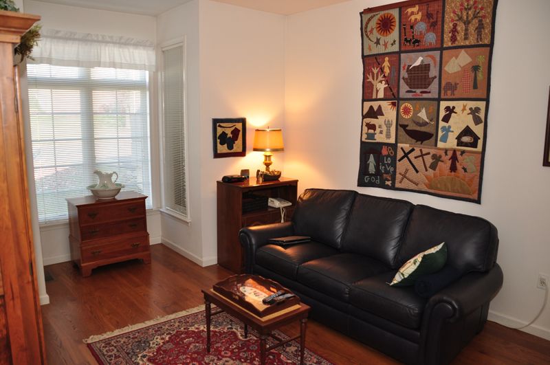 A cozy living room with a black leather couch, a decorative quilt on the wall, and a warm lamp on a side table.