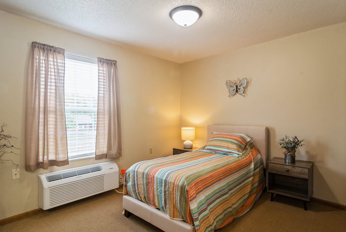 A cozy bedroom with a single bed covered in striped bedding, flanked by a nightstand and lamp near a window with curtains.