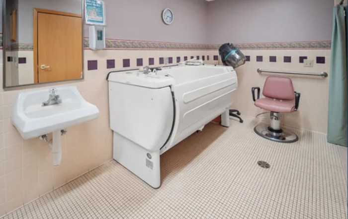 A healthcare facility room featuring a specialized bathtub and a salon chair equipped with a hair dryer.