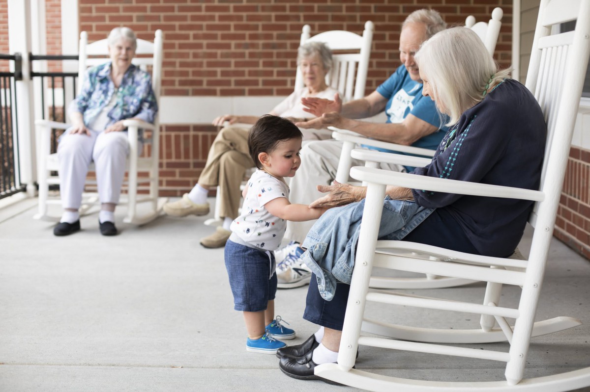 A young child interacts with elders on a porch filled with rocking chairs.