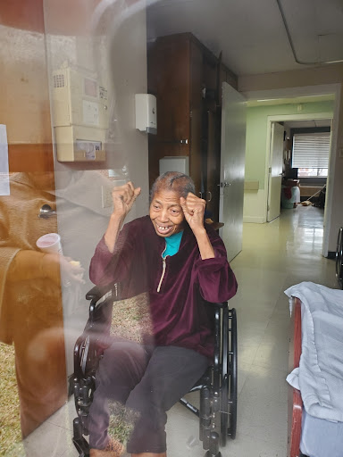 An elderly woman in a wheelchair joyfully gestures through a window, expressing happiness during a visit.