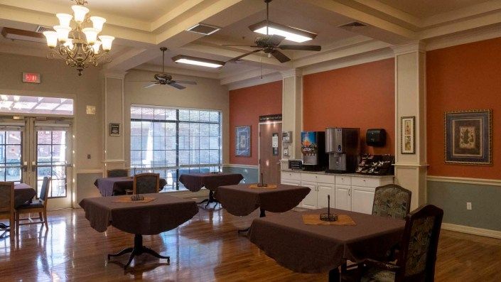 A cozy dining area with several tables and chairs, featuring a beverage station against the wall and large windows letting in natural light.