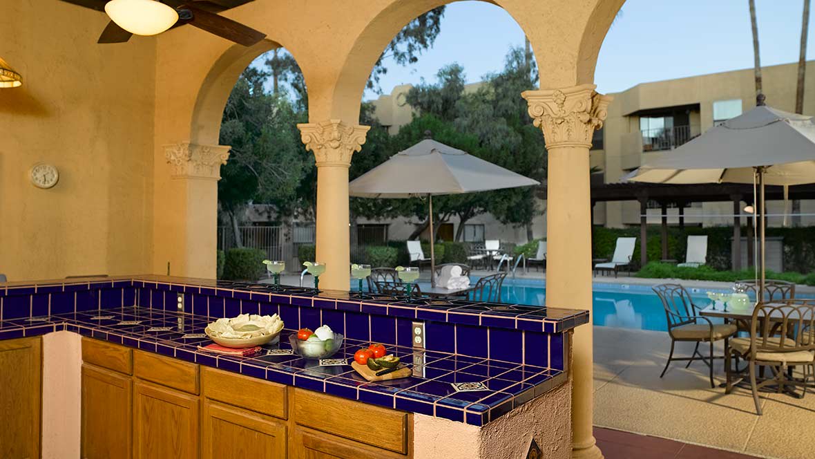 A Mediterranean-style outdoor kitchen with a blue tiled countertop overlooks a swimming pool and patio area.