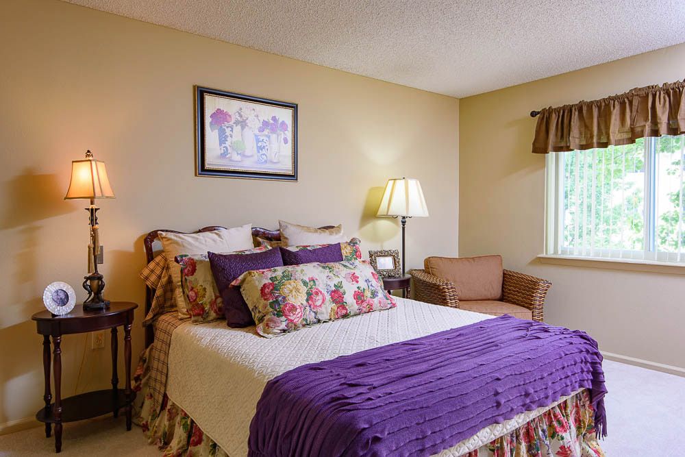 A cozy bedroom with vibrant purple accents, featuring a well-made bed, decorative pillows, a side lamp, and artwork on the wall.