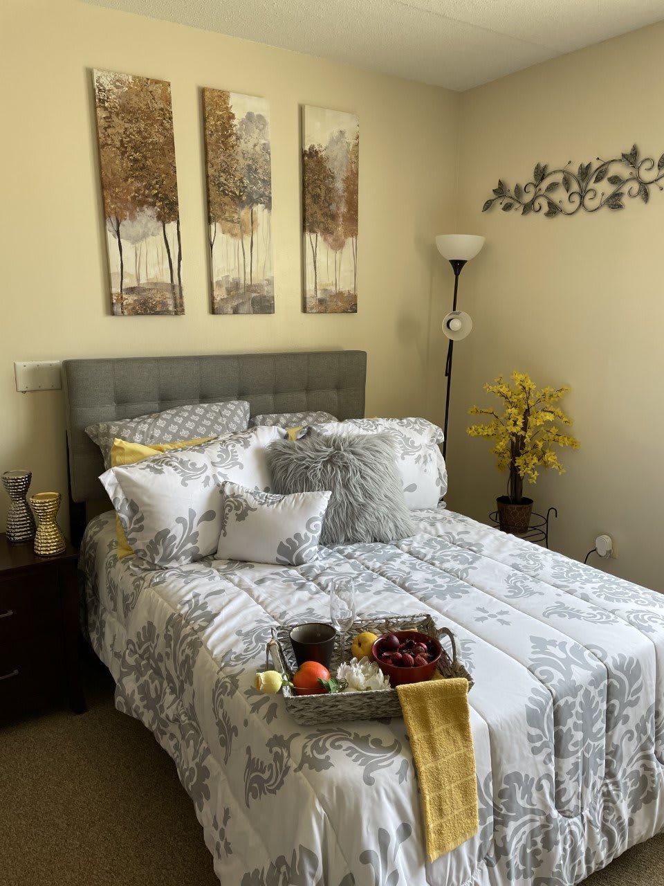 A cozy bedroom features a neatly made bed with patterned bedding, complemented by wall artwork and decorative elements like a floor lamp and plants. A tray with breakfast items is placed on the bed.