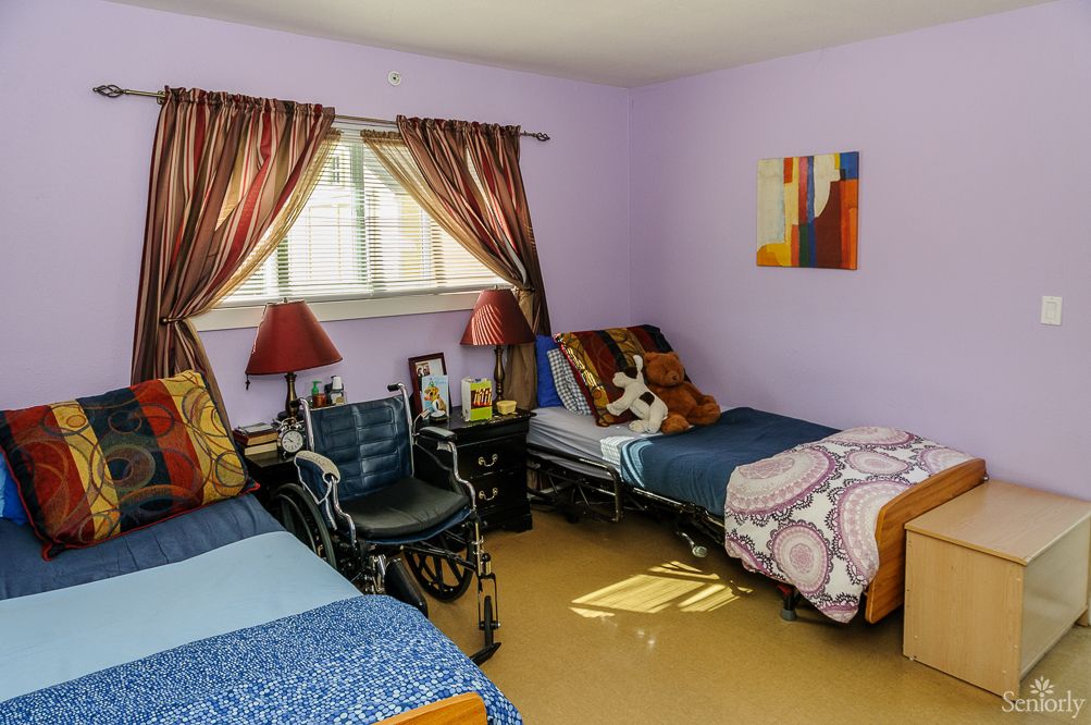 A cozy bedroom with twin beds, featuring a wheelchair between them and decorated with colorful curtains and a painting on the wall.