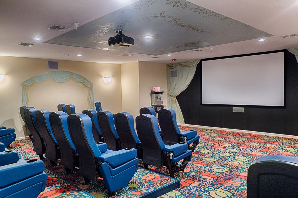 A home theater room with blue cinema-style seating and a large projection screen, featuring a vibrant carpet design.