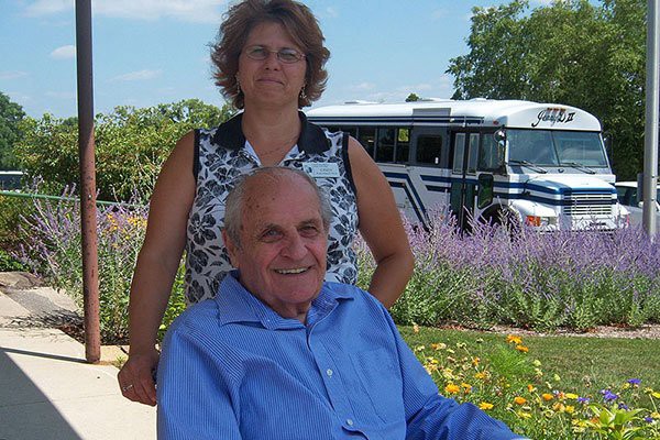 A smiling elderly man in a wheelchair sits in front of a woman with flowers and a bus in the background.
