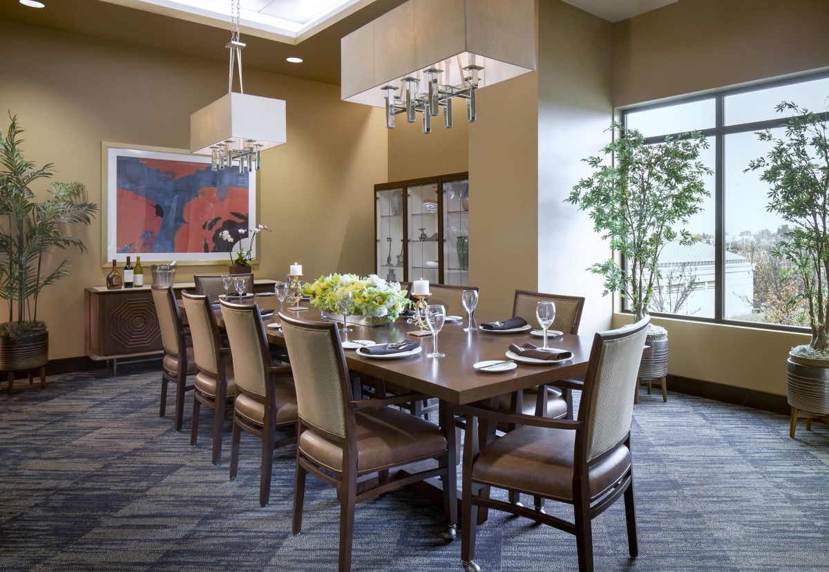A modern dining room with a large table and chairs, featuring contemporary artwork, chandeliers, and a large window.
