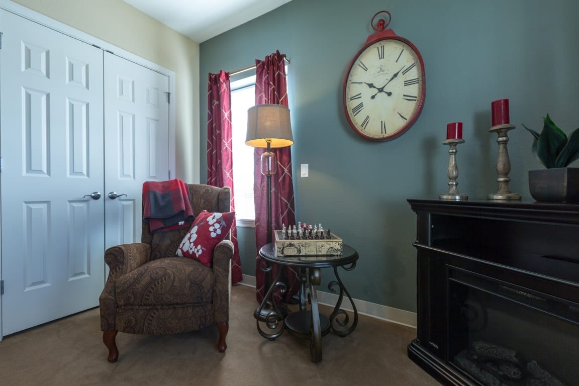 A cozy living room with an armchair and a side table topped with a chess set, illuminated by a lamp, featuring a large wall clock.