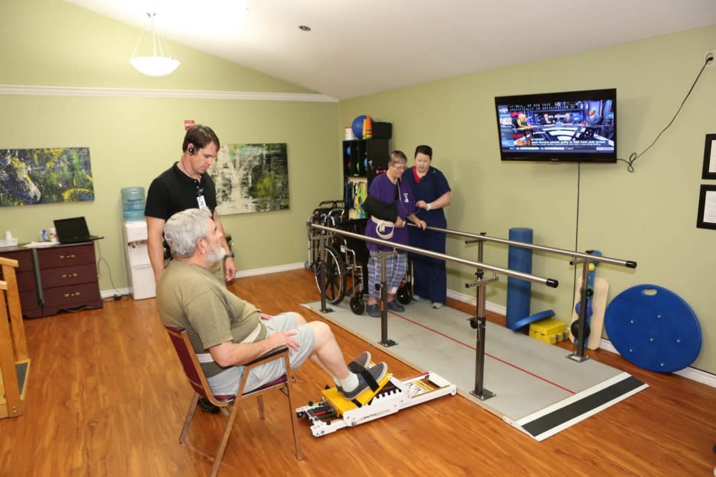 An elderly person participates in a physical therapy session with the assistance of medical staff in a treatment room equipped with exercise equipment.