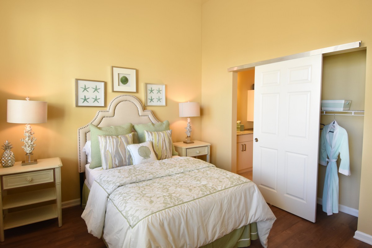 A cozy bedroom with pastel yellow walls, a neatly made bed with multiple pillows, two bedside tables with lamps, and decorative artwork on the walls.