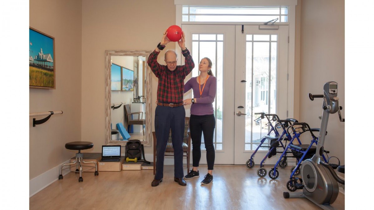 An elderly man participates in a physical therapy session, lifting a red exercise ball with the guidance of a therapist.