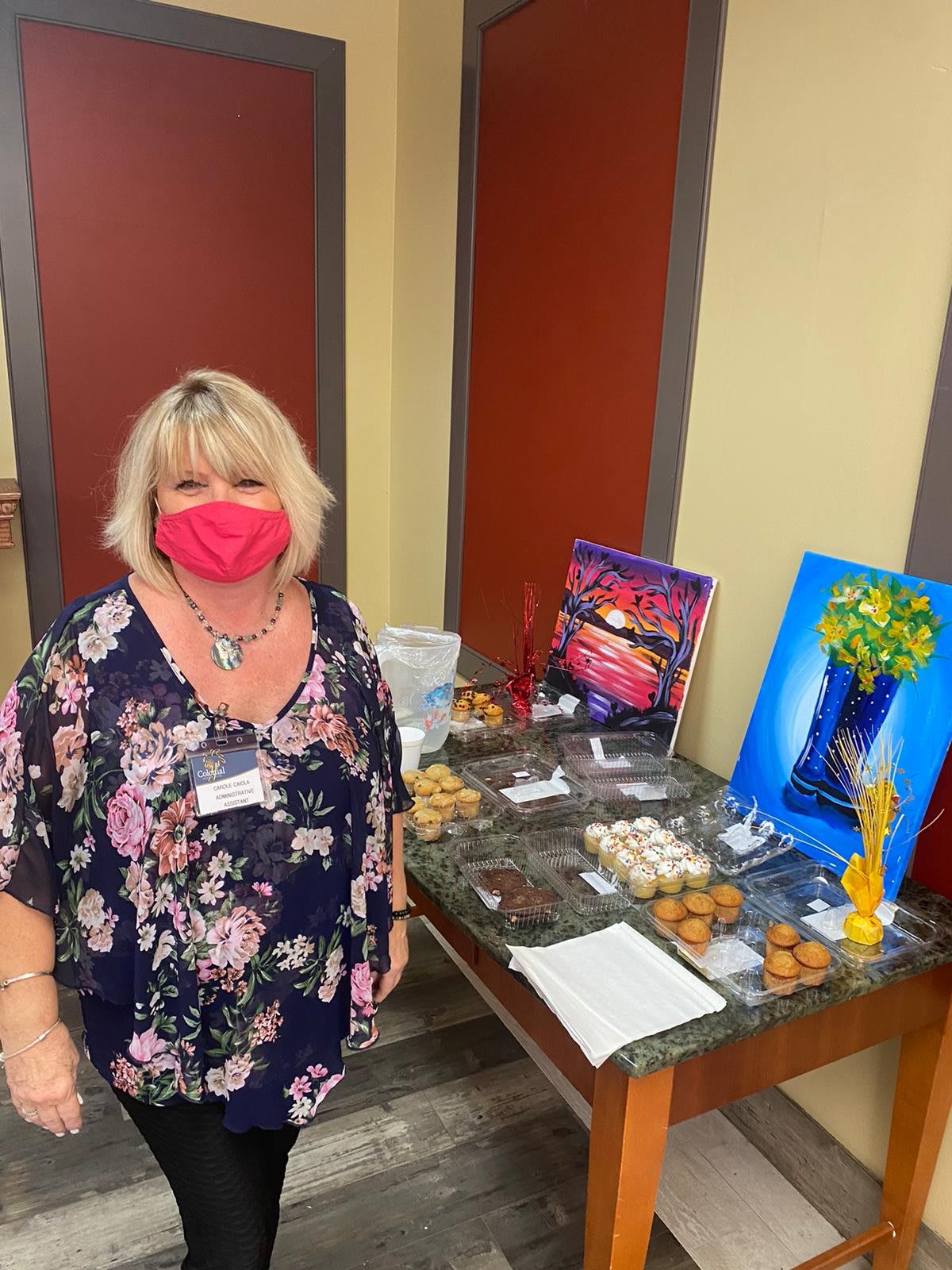 A woman wearing a floral shirt and a pink mask stands next to a table displaying cookies and artwork.