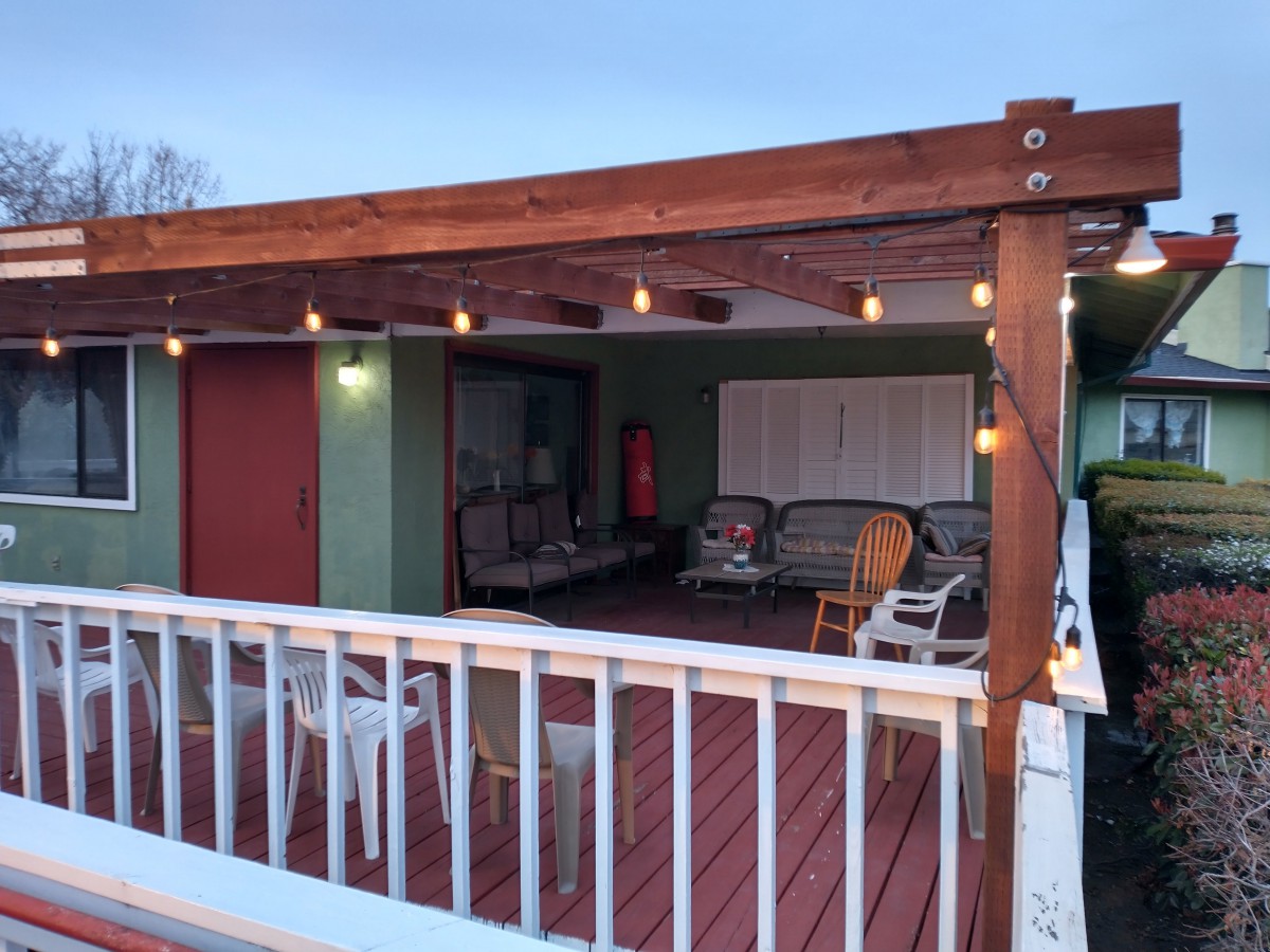 A cozy outdoor patio with a wooden deck and pergola, featuring string lights and comfortable seating.