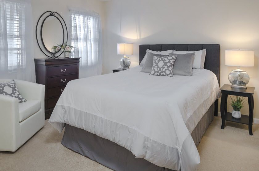 A neatly arranged bedroom featuring a bed with white linens, a lamp on the nightstand, and a dresser with a mirror.