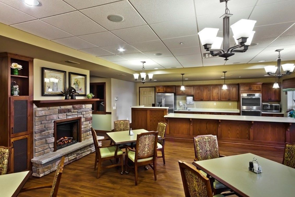 A cozy dining area with a fireplace and kitchen in the background, featuring tables and chairs under warm lighting.