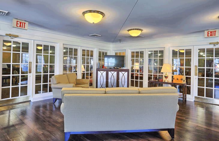 A cozy living room with sofas and chairs arranged around a central television, featuring large windows and warm lighting.
