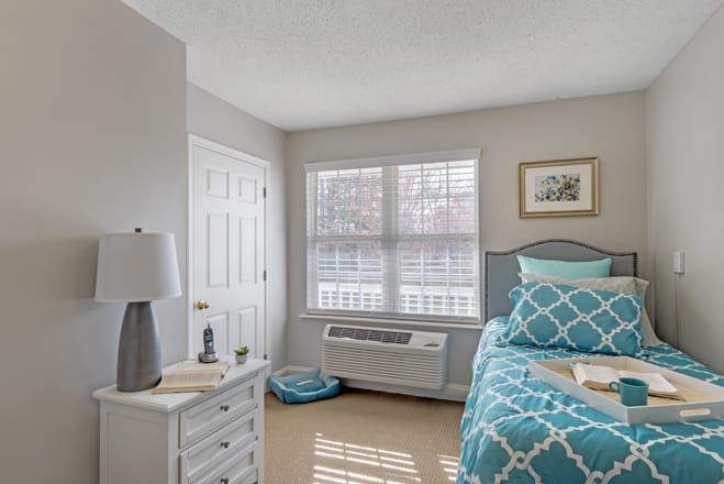 A cozy bedroom with a neatly made bed, bedside table, and a window letting in natural light.