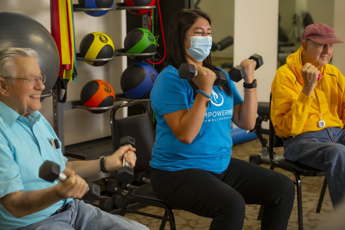 A group of people exercising with dumbbells in a gym setting, wearing masks for safety.