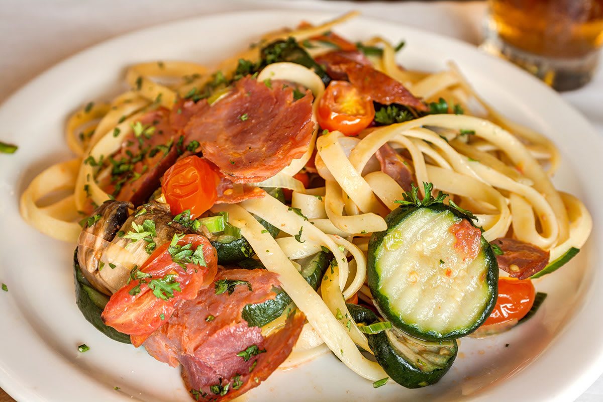 A plate of linguine pasta with zucchini, cherry tomatoes, and prosciutto garnished with fresh herbs.