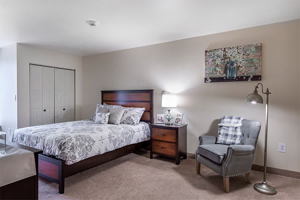 A cozy bedroom with a bed, nightstand, lamp, and a chair in neutral colors.