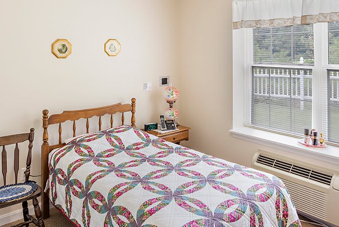 A cozy bedroom featuring a patterned quilt on the bed, with a nightstand and lamp by the window.