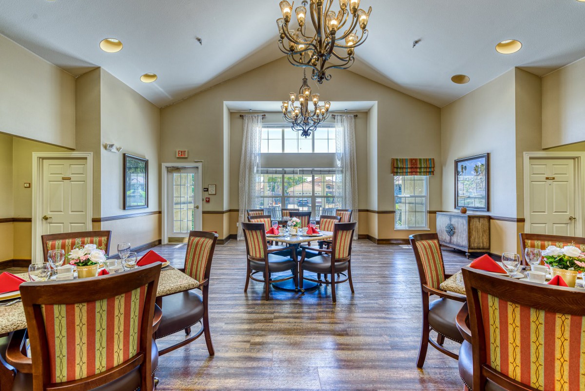 A well-lit dining room with wooden flooring and elegant chandeliers, featuring several tables and chairs arranged neatly.