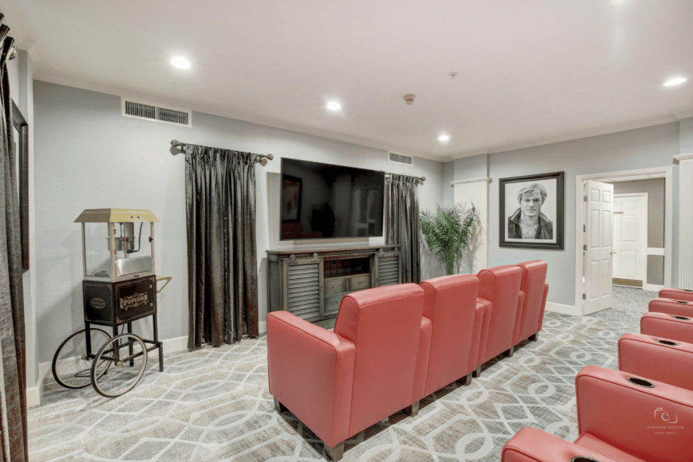 A cozy home theater room featuring red reclining chairs facing a large wall-mounted TV, with dark curtains and framed artwork on the walls.