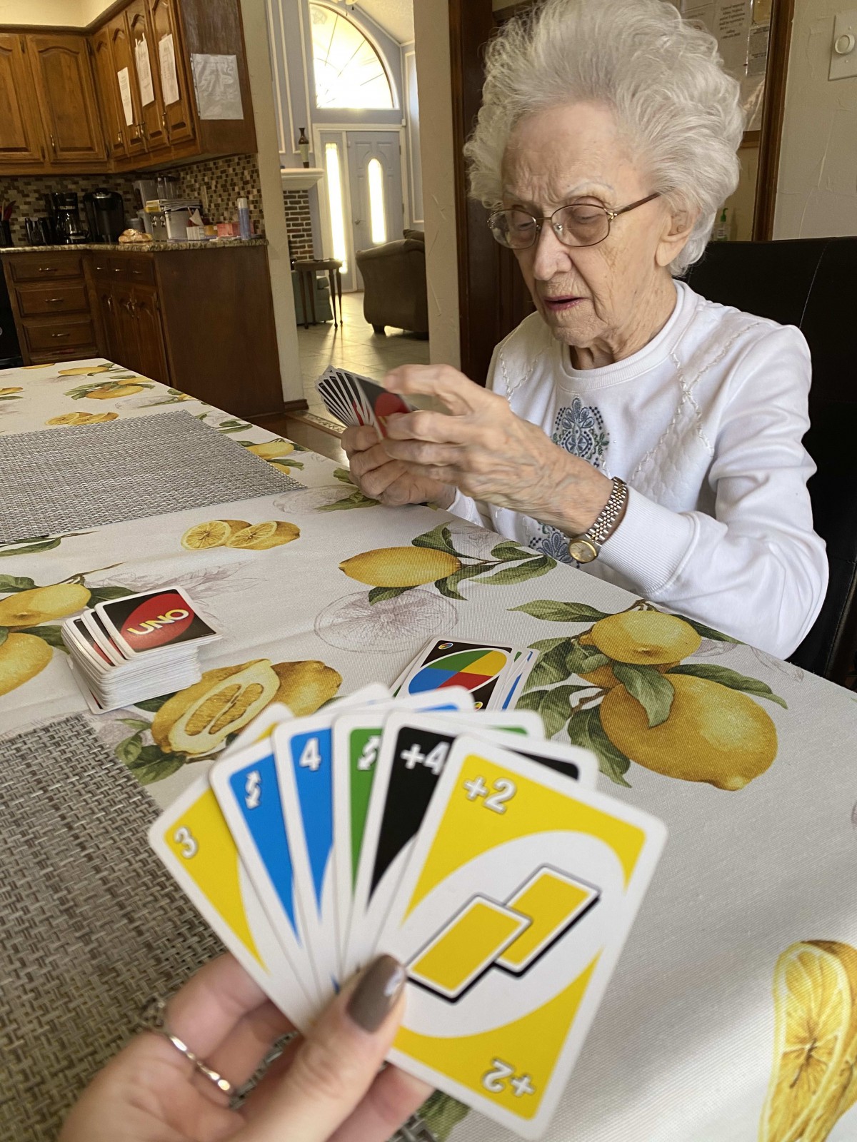 An elderly woman playing UNO at a table covered with a lemon-themed tablecloth.