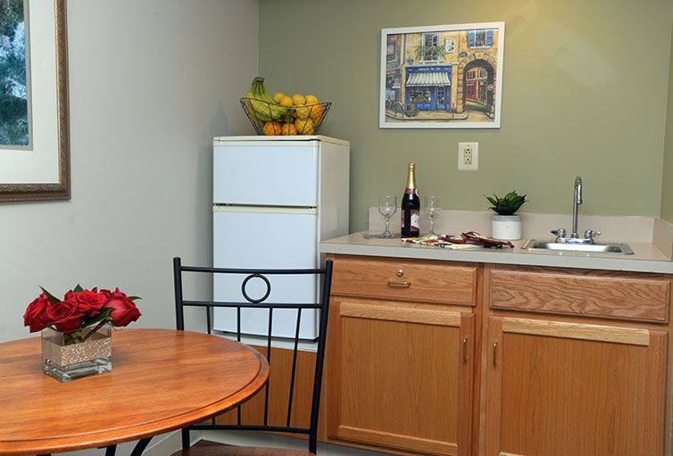 A cozy kitchen corner with a small table and chairs, featuring a fridge topped with a fruit basket, wooden cabinets, and a countertop adorned with a bottle and potted plant.