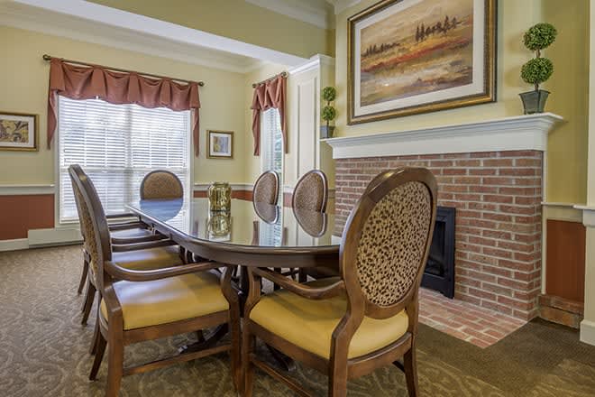 A cozy dining room with a glass table, upholstered chairs, and artwork above a brick fireplace.