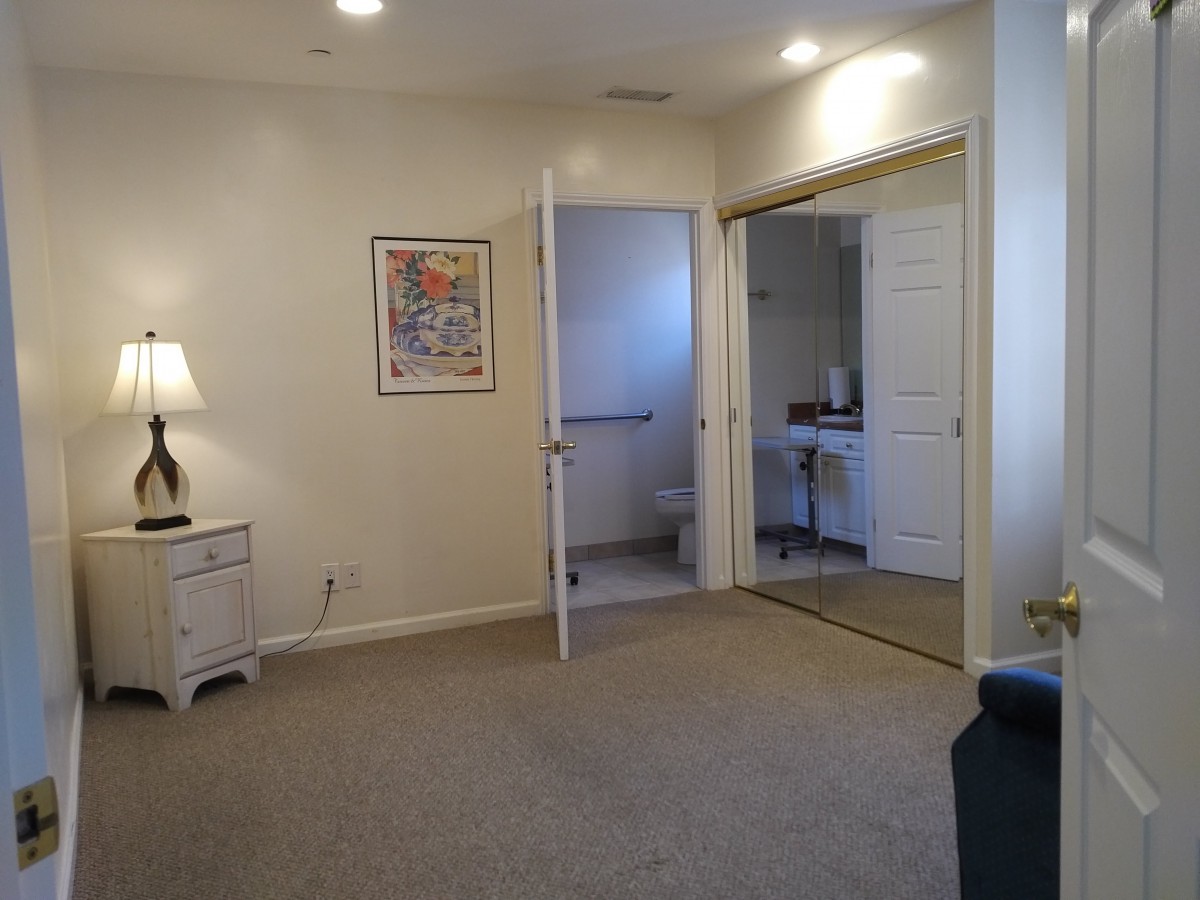 A spacious bedroom with neutral colors, featuring a lamp, a poster, an open door leading to a bathroom, and a closet.
