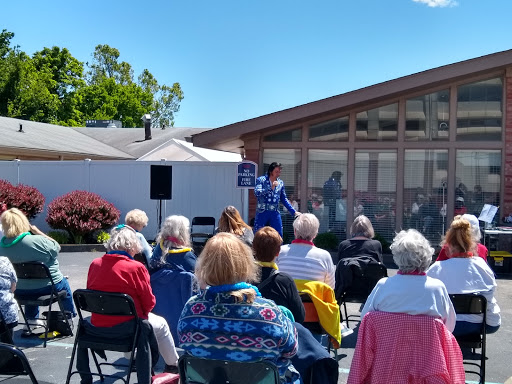 A community event taking place outdoors, with a speaker addressing a gathered audience of seniors.