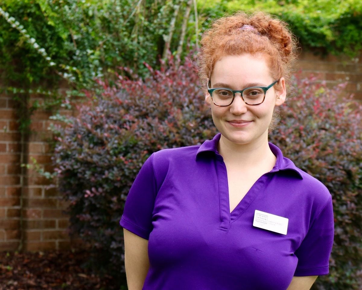 A person with red hair and glasses, wearing a purple shirt with a name tag, smiling in a garden.